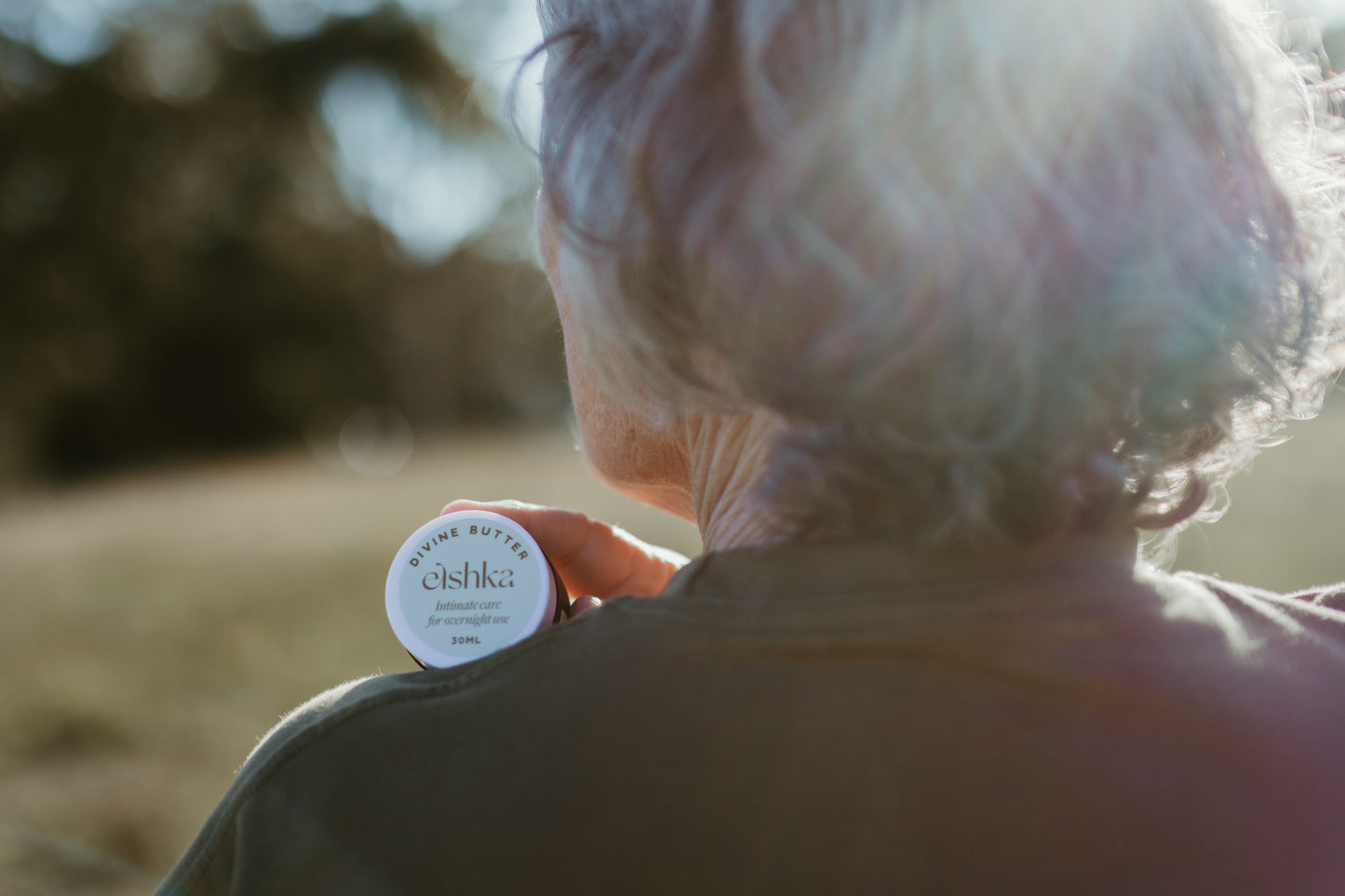An elderly woman viewed from behind in warm sunlight, holding a small round jar of Elshka Divine butter against her shoulder.
